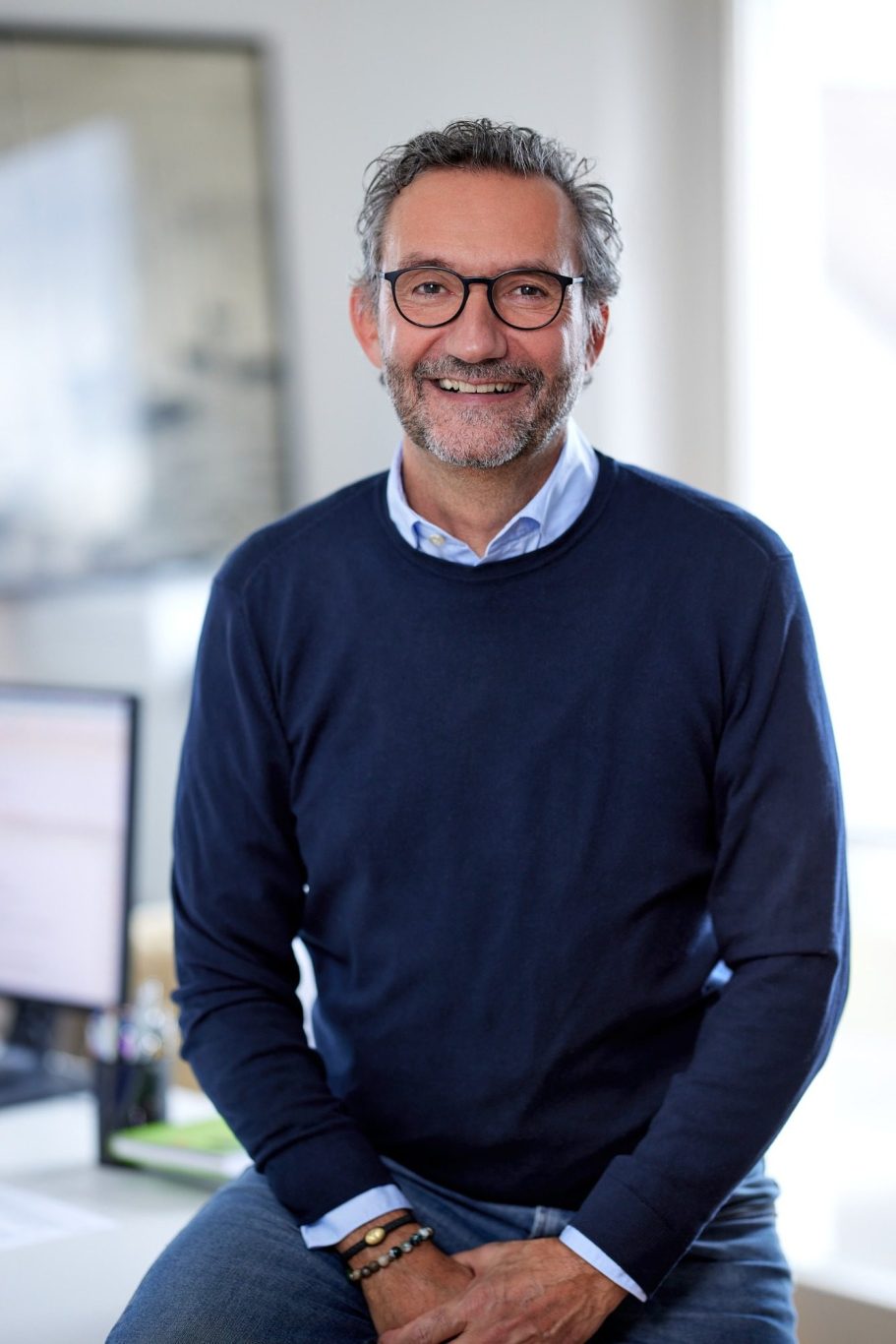 Mann mit Brille, lächelnd, in einem hellen Büro, sitzt auf einem Tisch.
