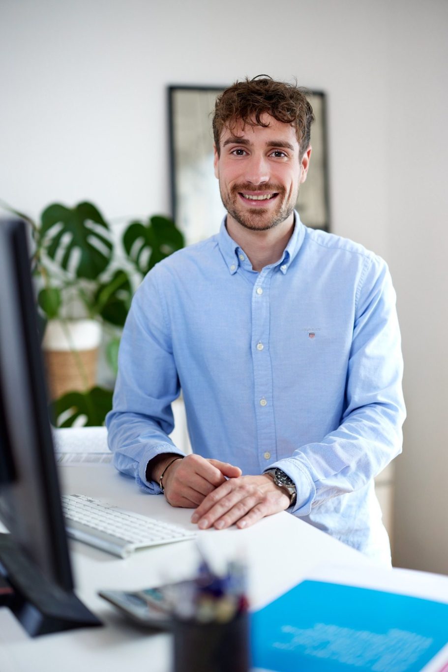 Junger Mann in blauer Hemd sitzt am Schreibtisch, lächelt freundlich.