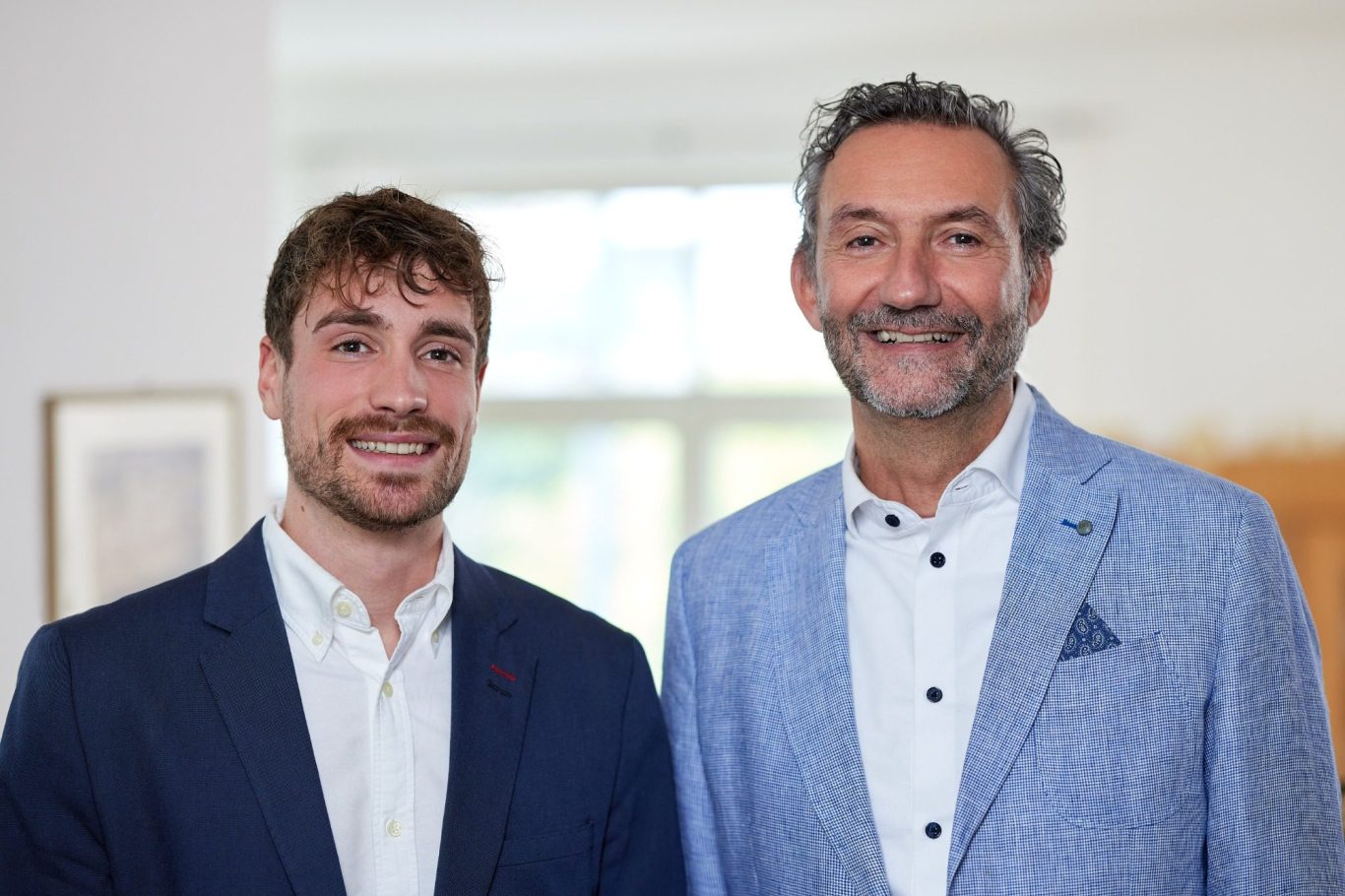 Zwei Männer stehen lächelnd nebeneinander, einer in einem dunklen Anzug, der andere in einem hellen Blazer.